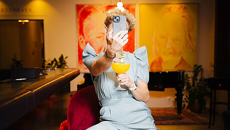 Person in light blue jumpsuit taking a selfie at bar with cocktail in hand