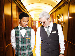 Two hotel staff members in uniform standing and smiling in a hallway with warm lighting