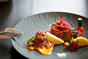 Beef tartare with pickled onions, potato wedges, cherry tomatoes, and hot sauce on a black plate