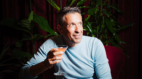 Mann im blauen Pullover mit Cocktail sitzt lächelnd in Lounge mit Zimmerpflanzen