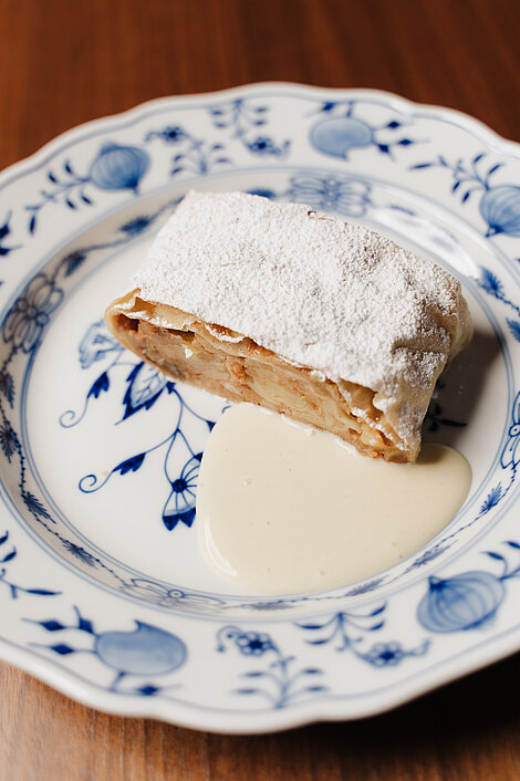 Slice of apple strudel with powdered sugar and vanilla sauce on floral porcelain plate