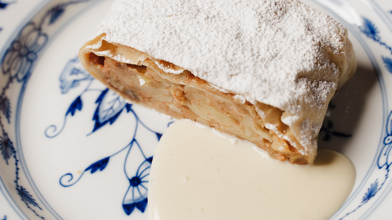 Slice of apple strudel with powdered sugar and vanilla sauce on floral porcelain plate