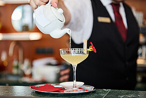 Bartender pouring cocktail from white teapot into coupe glass with red edible flower garnish