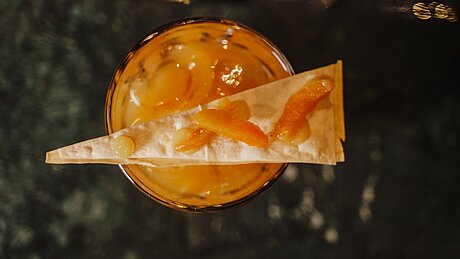 Orange cocktail with ice topped with a triangle crisp and candied orange peel