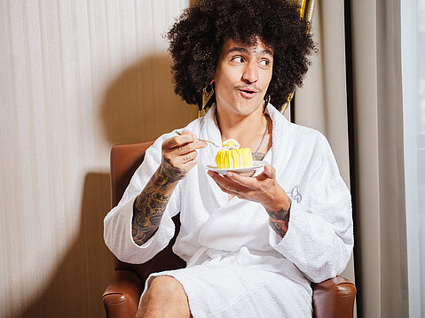 Person in white bathrobe sitting on chair enjoying dessert in hotel room