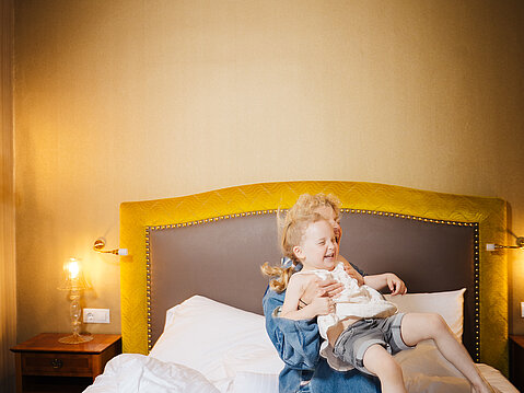 Happy child playing on a hotel bed with an adult in a cozy room setting