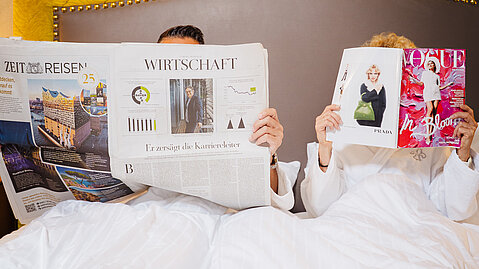 Paar im weißen Bademantel liest Zeitung und Modemagazin im Bett