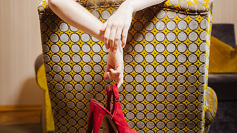 Woman resting over patterned armchair holding red high heel shoes